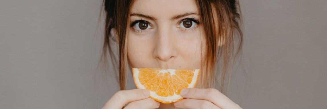 Close-up portrait of a woman holding a half orange slice, with a focus on natural beauty and skincare.